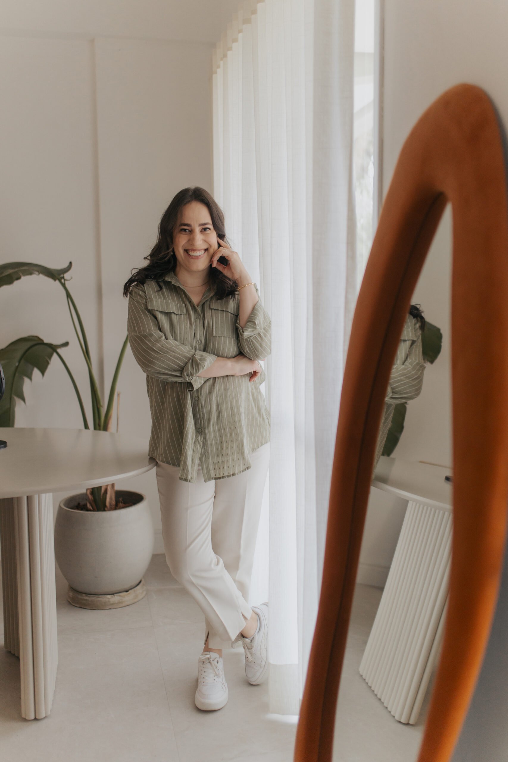 Mulher sorrindo, de cabelo escuro, usando blusa listrada em tom de verde, calça branca e tênis branco, posando na frente de um espelho de chão.