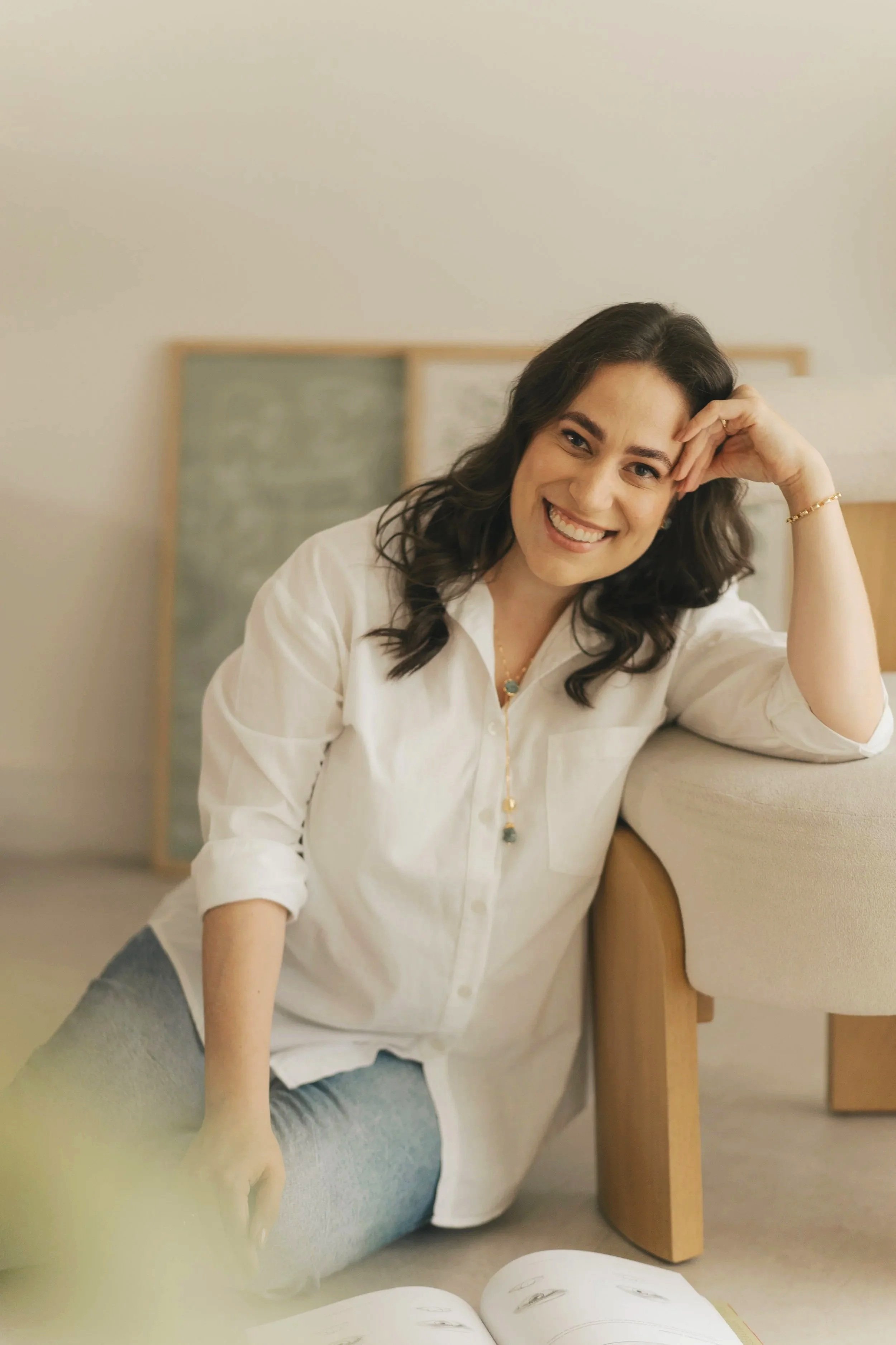Mulher de cabelo castanho, sorridente, sentada no chão, com braço apoiado no sofá, usando camisa branca e jeans, em ambiente interno confortável.